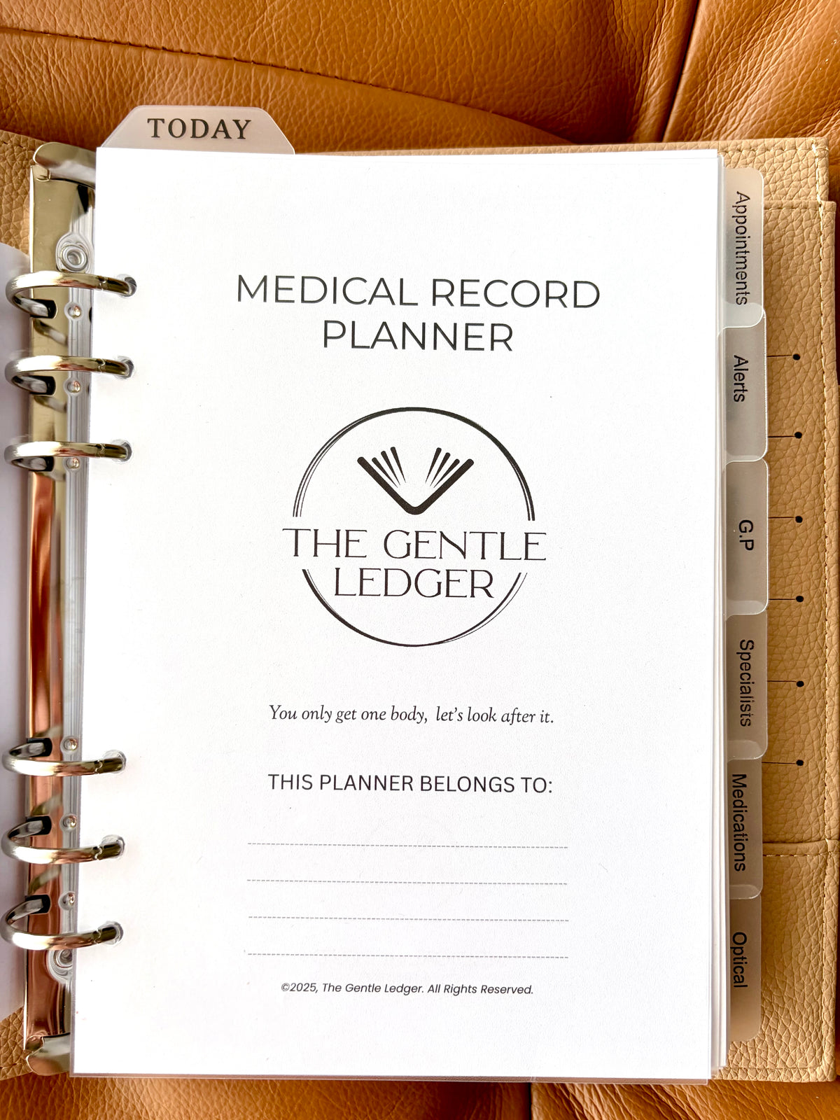 A5 ring binder Medical Record Planner from The Gentle Ledger, shown open on a tan leather surface. The white cover page features black text and a circular logo, with beige index tabs visible along the right side.