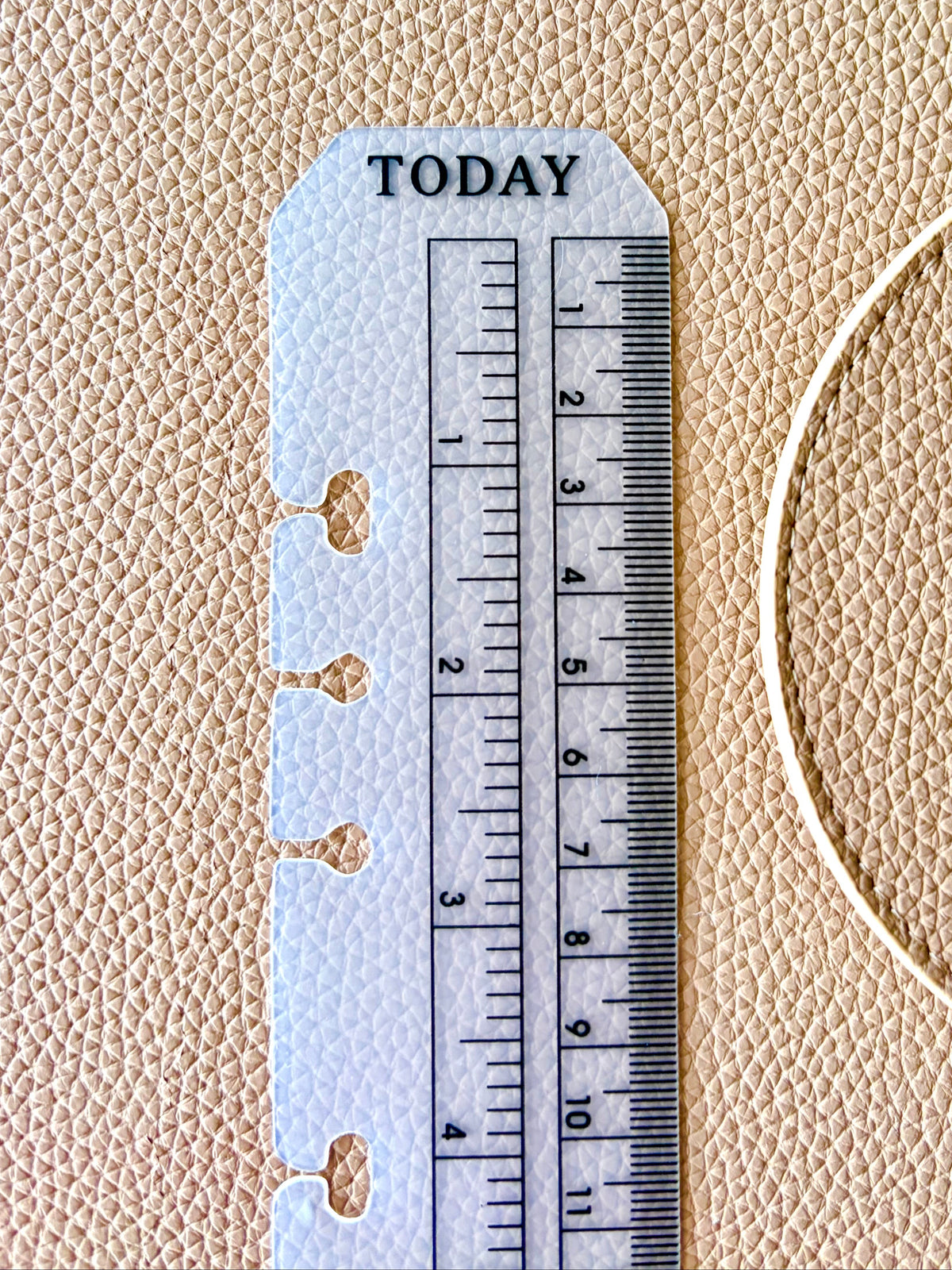 A white plastic planner insert with a ruler and 'TODAY' label, shown on a textured beige leather background next to a gold ring.